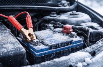Zamarznięty akumulator samochododowy w komorze silnika z widocznymi klemami i podłączonymi kablami rozruchowymi podczas zimowych warunków