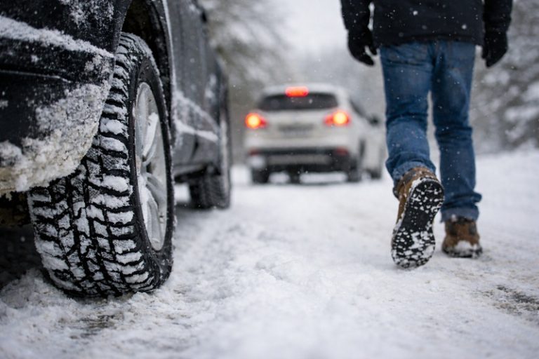Samochód na ośnieżonej drodze zimą, zbliżenie na oponę z bieżnikiem i kierowcę w zimowym obuwiu idącego w stronę auta podczas śnieżycy